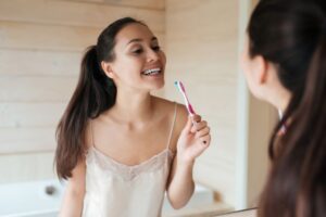 Woman holding her toothbrush, looking at her teeth in bathroom mirror